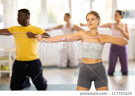 Girl conducts Pilates training for multinational students, standing exercises with wide elastic band 117205824