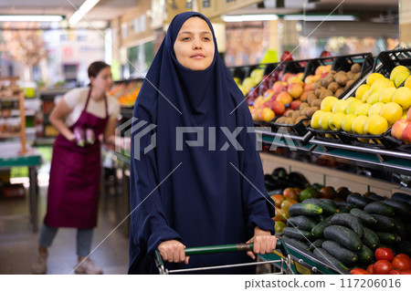 Muslim woman in dark blue hijab stands in grocery department of supermarket with grocery basket Muslim woman in dark blue hijab stands in grocery department of supermarket with grocery basket 117206016