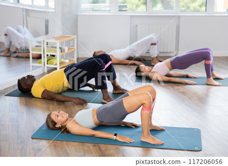 Girl performs glute bridge during Pilates multinational training with elastic band on shins Girl performs glute bridge during Pilates multinational training with elastic band on shins 117206056
