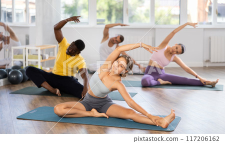 Young female teacher working out Parivritta Janu Shirshasana with multinational students group Young female teacher working out Parivritta Janu Shirshasana with multinational students group 117206162