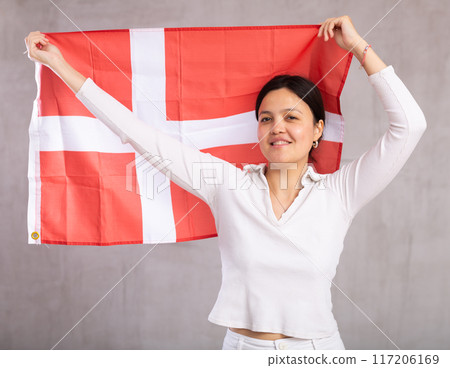 Middle-aged woman in casual clothes holds unfurled flag of Denmark in hands raised above head 117206169