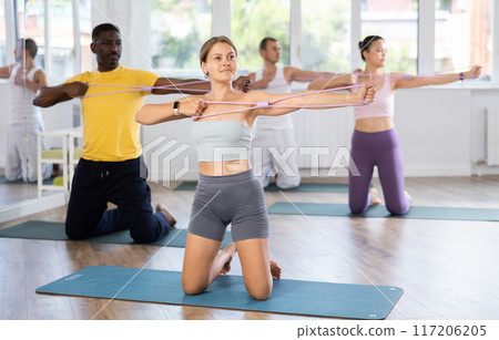 Young woman performs exercises with resistance band during multinational Pilates training, kneeling Young woman performs exercises with resistance band during multinational Pilates training, kneeling 117206205