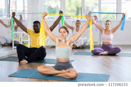 Group of multinational women and men sitting on sports mats and doing arm stretches using resistance bands at Pilates studio Group of multinational women and men sitting on sports mats and doing arm stretches using resistance bands at Pilates studio 117206231