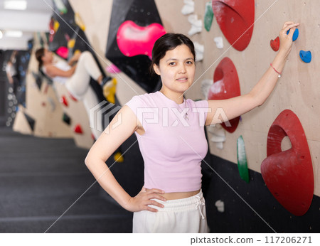 Young smiling Asian girl coach stands at climbing wall and leans holding on to hook. Young smiling Asian girl coach stands at climbing wall and leans holding on to hook. 117206271