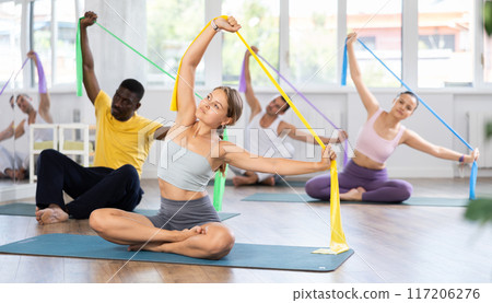Girl conducts Pilates multinational group sitting with elastic band in hands above head body bends Girl conducts Pilates multinational group sitting with elastic band in hands above head body bends 117206276