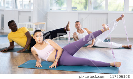Pilates class - group of people of different nationalities doing leg stretches using resistance bands in gym Pilates class - group of people of different nationalities doing leg stretches using resistance bands in gym 117206368
