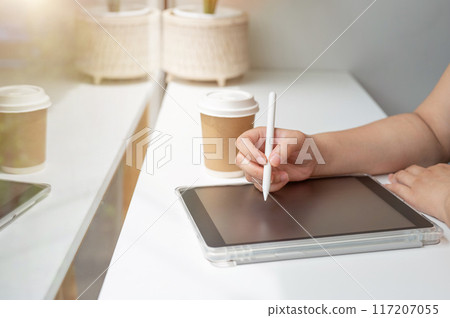 A woman holding a stylus pen, writing on a digital tablet, working remotely from a coffee shop. 117207055