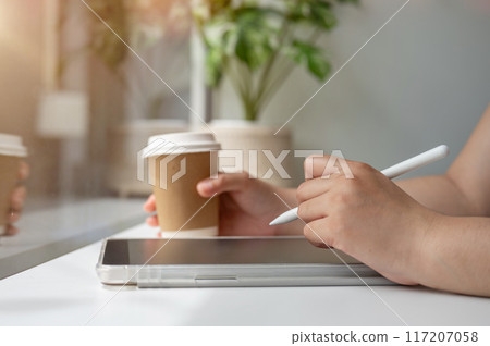 A woman holding a stylus pen, writing on a digital tablet, working remotely from a coffee shop. 117207058