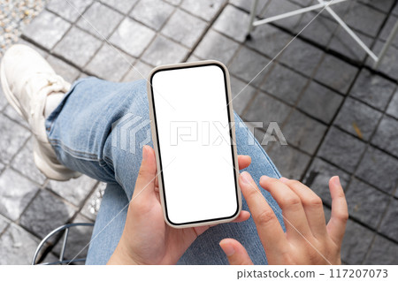 Top view image of a woman in jeans sitting outdoors, using her smartphone, touching on the screen. Top view image of a woman in jeans sitting outdoors, using her smartphone, touching on the screen. 117207073