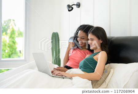 Happy young couple enjoy hotel room or house bedroom together. Two beautiful multiethnic women smiling and surfing the web on laptop in bed.  Lesbian couple together indoors concept. 117208904