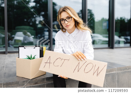 Woman sitting outdoor with poster Act Now next to box of stuff . Beautiful female protester at strike against unemployment Woman sitting outdoor with poster Act Now next to box of stuff . Beautiful female protester at strike against unemployment 117209593