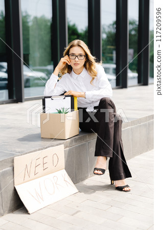 Woman sitting outdoor with cardboard banner with the slogan Need work next to box of stuff. Beautiful female protester at strike against unemployment 117209596
