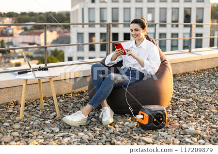 Woman relaxing on rooftop beanbag chair using smartphone and portable charger Woman relaxing on rooftop beanbag chair using smartphone and portable charger 117209879