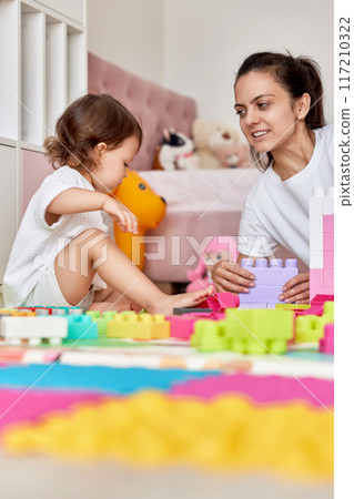 cute baby girl and her loving mother build tower from colorful cubes, having fun together 117210322