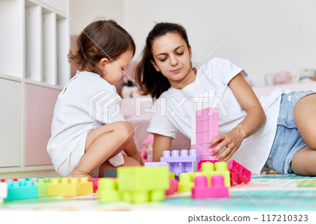 cute baby girl and her loving mother playing with colorful blocks in the children room, having fun together 117210323