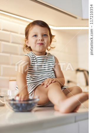 Little baby girl eating fresh blueberries from glass bowl in white kitchen Little baby girl eating fresh blueberries from glass bowl in white kitchen 117210354