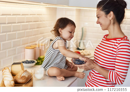 Little baby girl with her mother eating fresh blueberries in white kitchen Little baby girl with her mother eating fresh blueberries in white kitchen 117210355