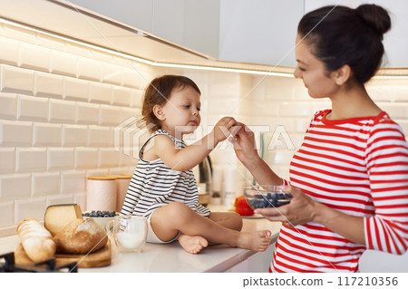 Little baby girl with her mother eating fresh blueberries in white kitchen Little baby girl with her mother eating fresh blueberries in white kitchen 117210356