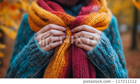 Hands tying a cozy knitted scarf in warm autumn colors with a fall landscape in blurred background 117211112