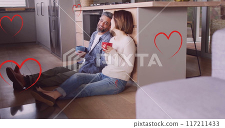 Image of hearts over diverse couple drinking coffe and sitting on floor in kitchen at home 117211433