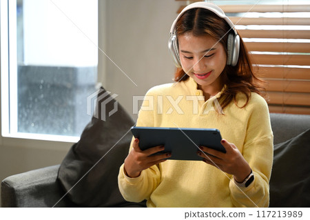 Pretty young Asian woman wearing headphones watching videos on digital tablet at home Pretty young Asian woman wearing headphones watching videos on digital tablet at home 117213989