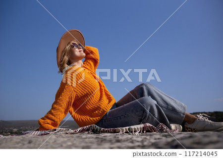 A woman in an orange sweater and blue jeans is sitting on a blanket on a rock A woman in an orange sweater and blue jeans is sitting on a blanket on a rock 117214045