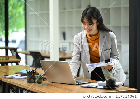 Shot of asian woman office worker holding documents and using laptop at desk Shot of asian woman office worker holding documents and using laptop at desk 117214061