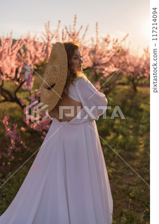 Woman blooming peach orchard. Against the backdrop of a picturesque peach orchard, a woman in a long white dress and hat enjoys a peaceful walk in the park, surrounded by the beauty of nature. Woman blooming peach orchard. Against the backdrop of a picturesque peach orchard, a woman in a long white dress and hat enjoys a peaceful walk in the park, surrounded by the beauty of nature. 117214094