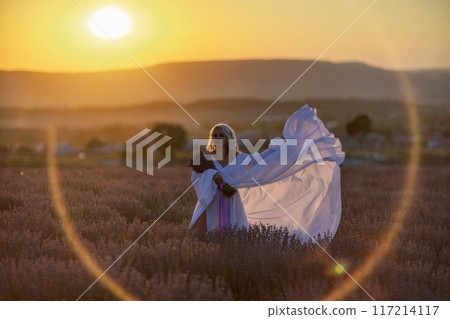Blonde woman poses in lavender field at sunset. Happy woman in white dress holds lavender bouquet. Aromatherapy concept, lavender oil, photo session in lavender 117214117