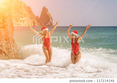 Women Santa hats ocean play. Seaside, beach daytime, enjoying beach fun. Two women in red swimsuits and Santa hats are enjoying themselves in the ocean waves. 117214148