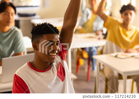 In high school, smiling teenage boy raising hand in classroom with classmates In high school, smiling teenage boy raising hand in classroom with classmates 117214210