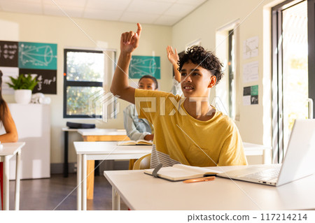 In high school, teenage boy raising hand while sitting at desk with laptop In high school, teenage boy raising hand while sitting at desk with laptop 117214214