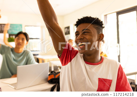 In high school, smiling teenage boy raising hand in classroom, participating actively In high school, smiling teenage boy raising hand in classroom, participating actively 117214218