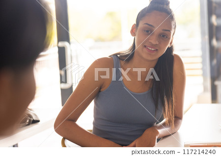 Teenager smiling and sitting at desk in high school classroom, engaging with friend 117214240