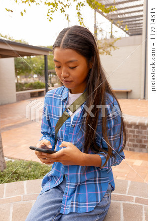 Texting on smartphone, teenage girl sitting outdoors at high school during break 117214315