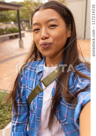 Teenage girl taking selfie outdoors, wearing blue plaid shirt, smiling confidently 117214316