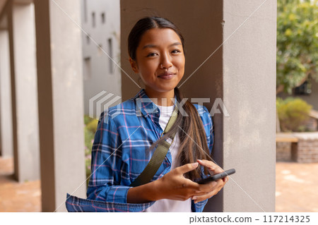 Holding smartphone and smiling, teenager standing outside high school building 117214325