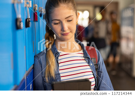 At high school, teenage girl using tablet while leaning against lockers, smiling 117214344