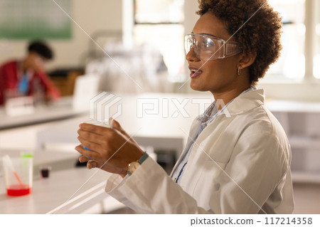 In high school, teacher wearing lab coat and safety goggles holding beaker in classroom 117214358