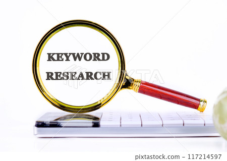 KEYWORD RESEARCH written conceptual phrase through a magnifying glass on a calculator on a light background 117214597