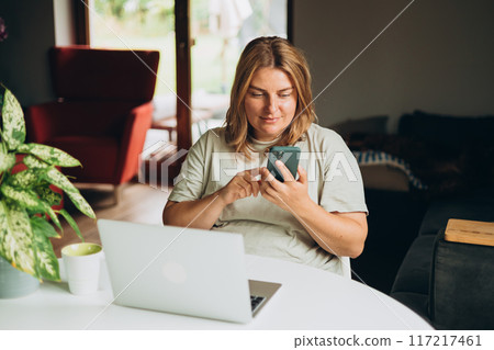 Happy 30s woman doing online shopping over laptop with phone at desk in home office. One young Caucasian woman is buying online on her laptop indoors 117217461