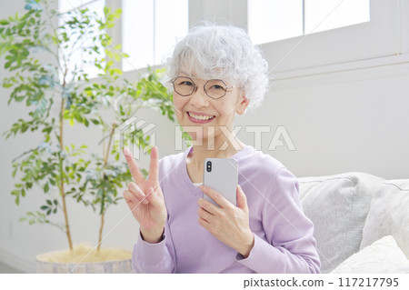 Senior woman making a peace sign with a smartphone in the living room Senior woman making a peace sign with a smartphone in the living room 117217795