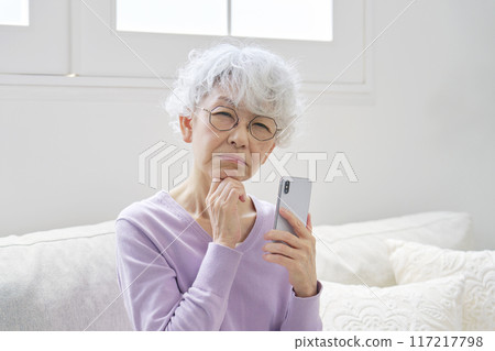 Senior woman worried about her smartphone in living room, looking at camera 117217798