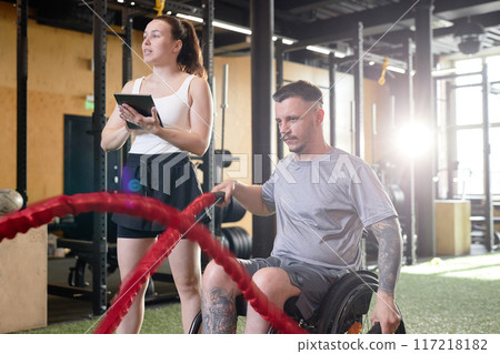Side view of man with disability using wheelchair during strength training in gym with female coach assisting, copy space Side view of man with disability using wheelchair during strength training in gym with female coach assisting, copy space 117218182