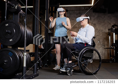 Full length portrait of woman with disability using VR technology in gym with female coach copy space 117218203
