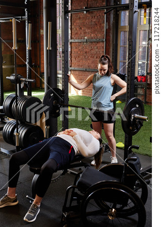 Vertical full length shot of woman with disability lying on bench during strength workout in gym with coach assisting Vertical full length shot of woman with disability lying on bench during strength workout in gym with coach assisting 117218244