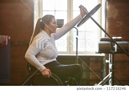 Side view portrait of adult woman with disability enjoying weight training with barbell in hazy gym Side view portrait of adult woman with disability enjoying weight training with barbell in hazy gym 117218246