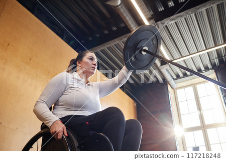 Low angle view of woman with disability using wheelchair and doing exercises with barbell during strength workout in gym copy space Low angle view of woman with disability using wheelchair and doing exercises with barbell during strength workout in gym copy space 117218248