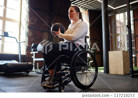 Dramatic side view of active woman using wheelchair in gym and doing strength training with contouring sunlight copy space 117218253