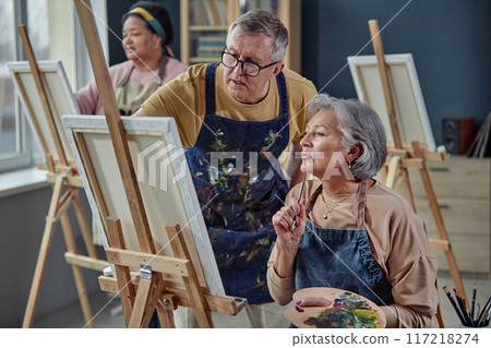 Portrait of two creative senior people painting picture on easel in art studio with senior man giving comments during art class copy space Portrait of two creative senior people painting picture on easel in art studio with senior man giving comments during art class copy space 117218274